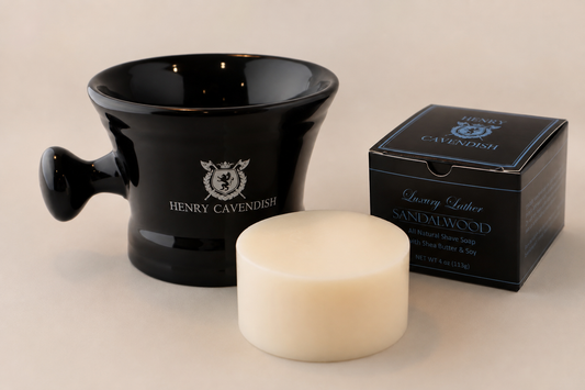 Henry Cavendishi Sandalwood Shaving Soap bar shown in front of its black packaging box and a black ceramic shaving bowl all on a neutral beige background.