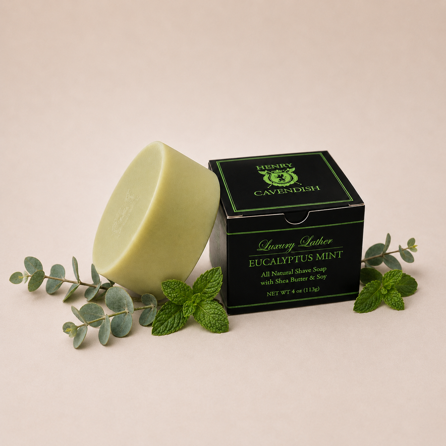 A bar of eucalyptus mint luxury shaving soap stands next to its black and green Henry Cavendish brand box, styled with fresh mint leaves and eucalyptus branches on a warm beige background.