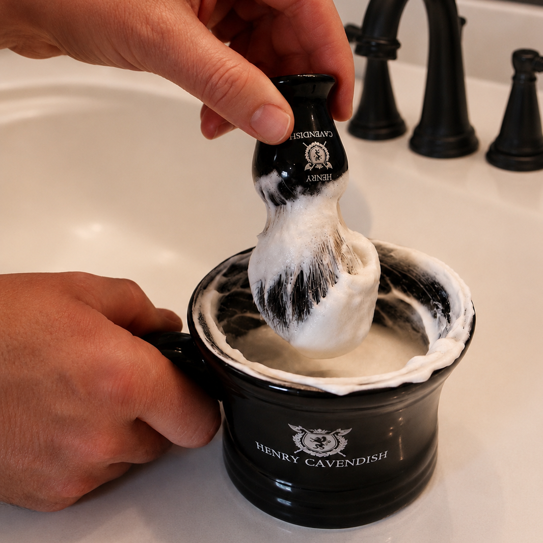 A hand holds a black shaving brush covered in white lather above a black shaving bowl on a white sink, with a black faucet in the background. The bowl and brush are branded Henry Cavendish.