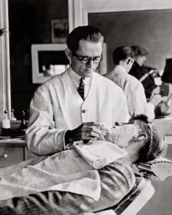 Black and white photograph of a man in a white lab coat performing a facial treatment on a reclining customer in a vintage barbershop or clinic setting.