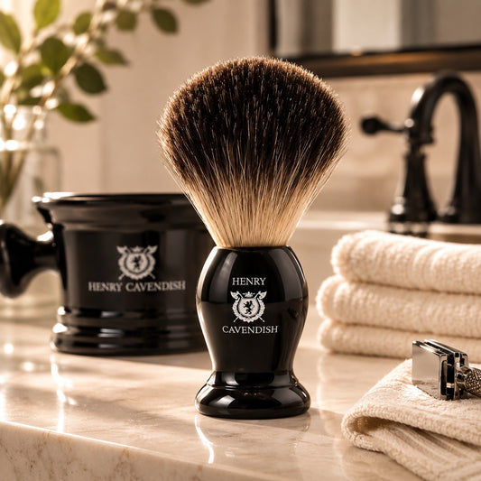 A black shaving brush with a large brown and cream bristle head stands on a marble bathroom counter, with a matching Henry Cavendish black shaving bowl, folded towels, and a razor in the background.