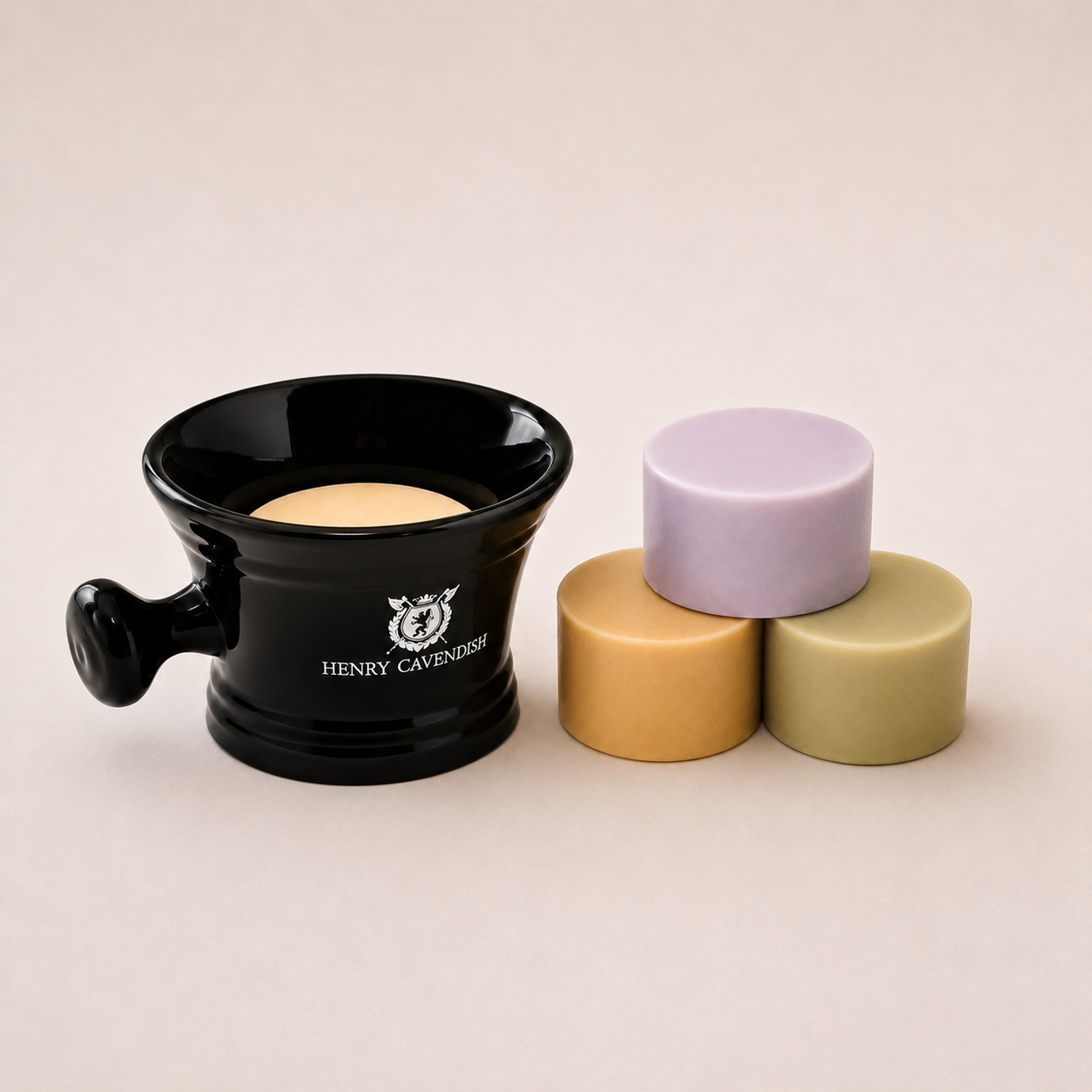 A black ceramic candle holder with a handle and a visible candle, alongside three small cylindrical candles in lavender, tan, and olive green, on a warm beige background. The holder is branded 'HENRY CAVENESSI'.
