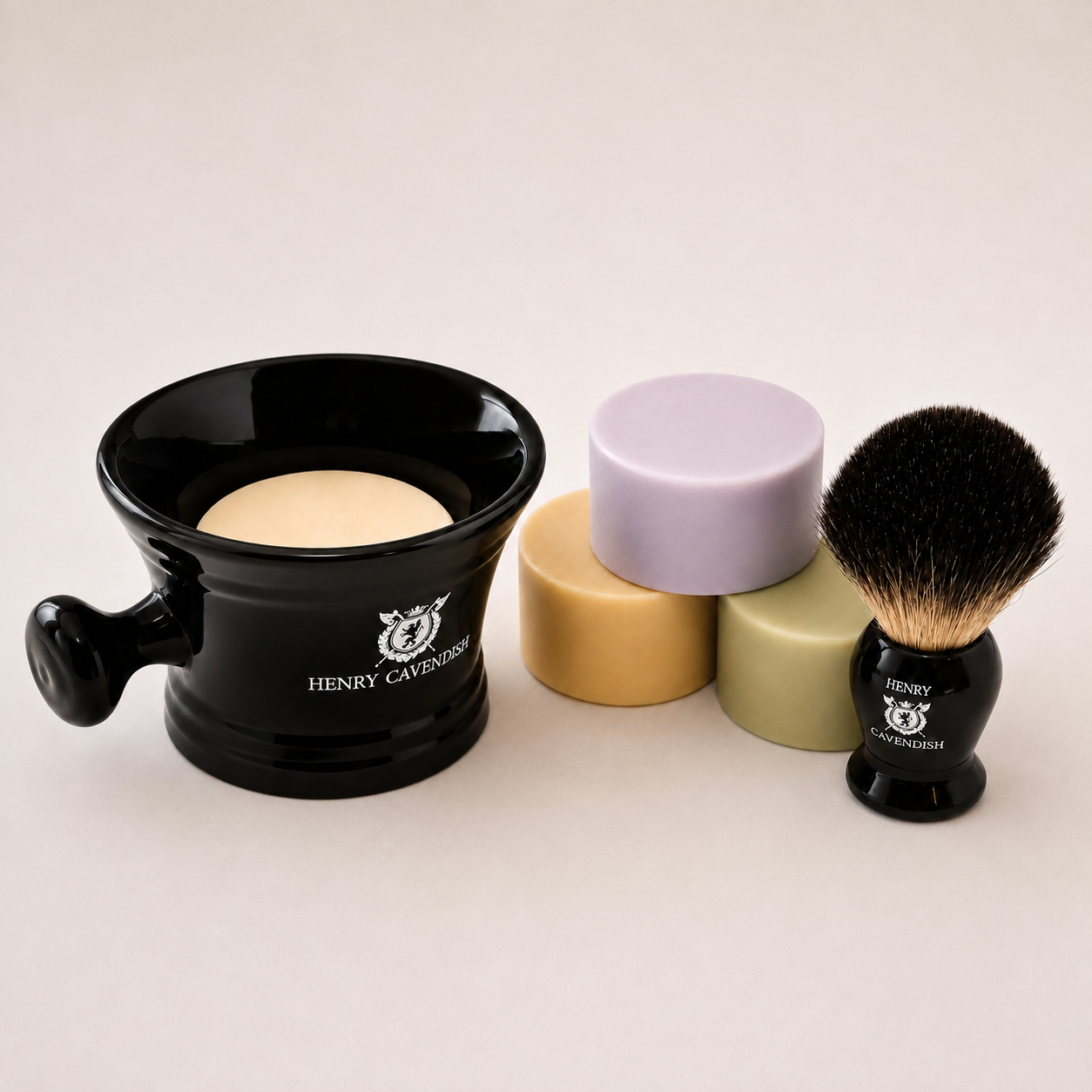 A shaving set by Henry Cavington on a light beige background, featuring a black ceramic shaving bowl with a beige soap puck, two pastel shaving stones in lavender and olive green, and a black shaving brush with black and beige bristles.