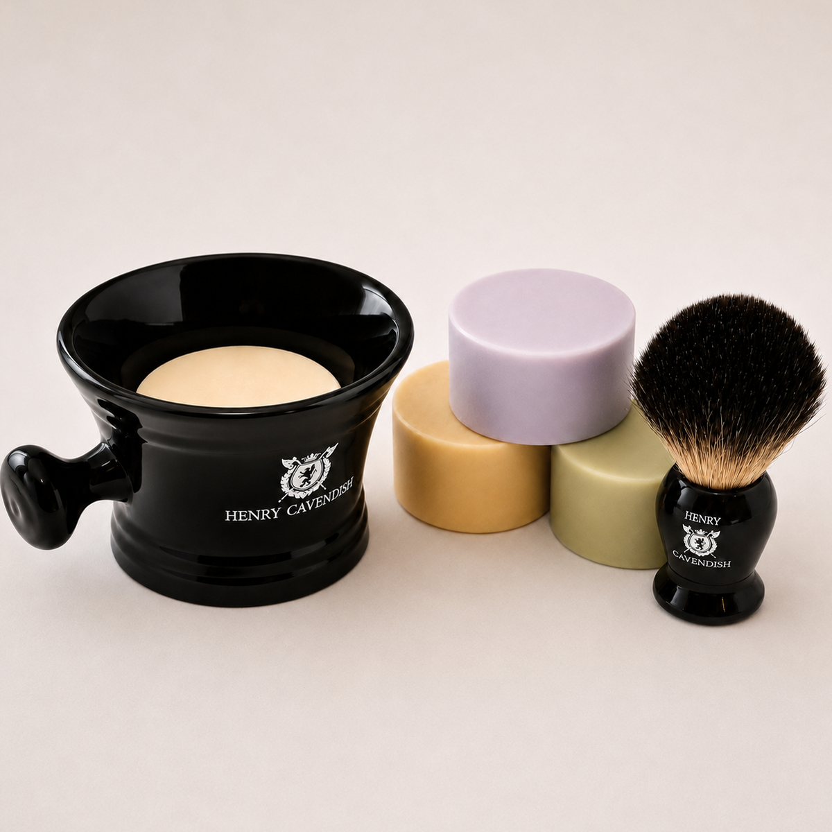 A shaving set by Henry Cavington on a light beige background, featuring a black ceramic shaving bowl with a beige soap puck, two pastel shaving stones in lavender and olive green, and a black shaving brush with black and beige bristles.