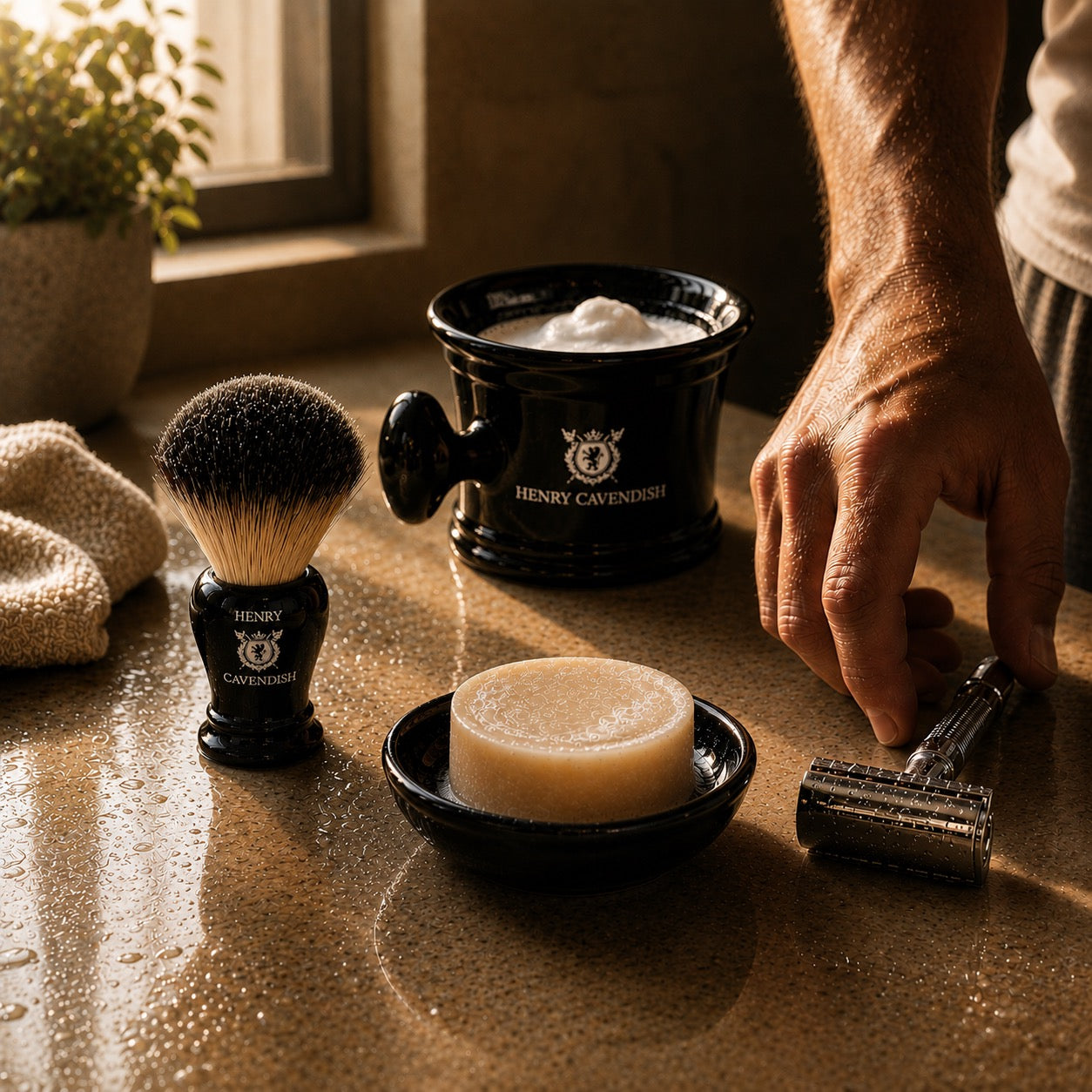 A close-up of a shaving setup on a wet stone countertop, featuring a black Henry Cavendish shaving brush, black shaving bowl with lather, a bar of soap in a black holder, and a metal double-edge razor being held by a person's hand.